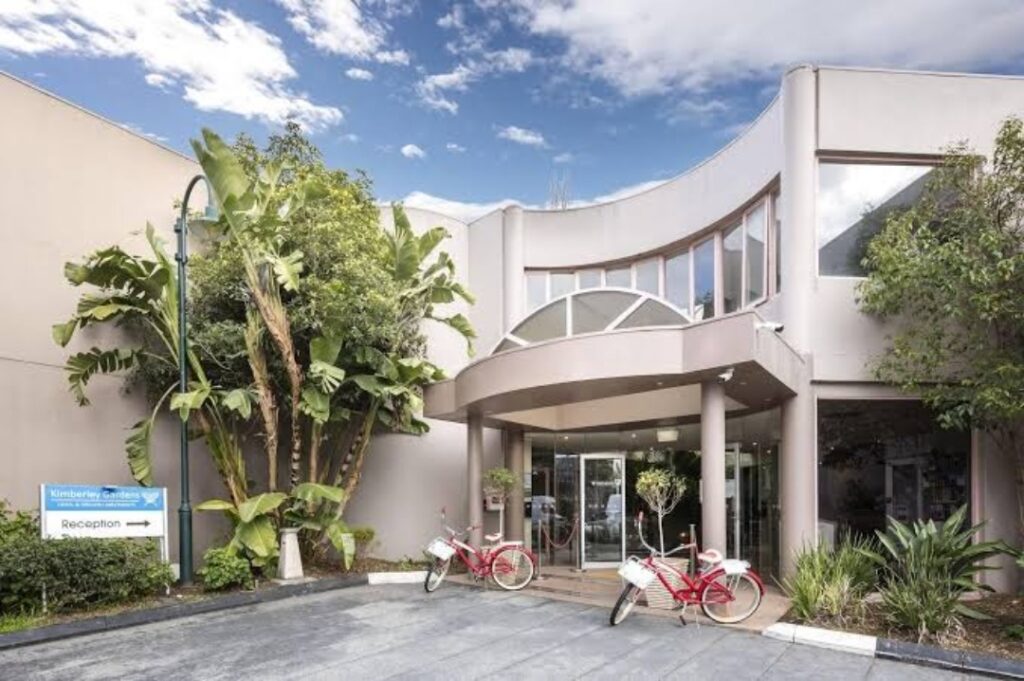 Front entrance of Kimberley Gardens Hotel at 441 Inkerman Street, St Kilda East, Melbourne, featuring a modern curved facade, tropical landscaping, and red bicycles near the reception area.