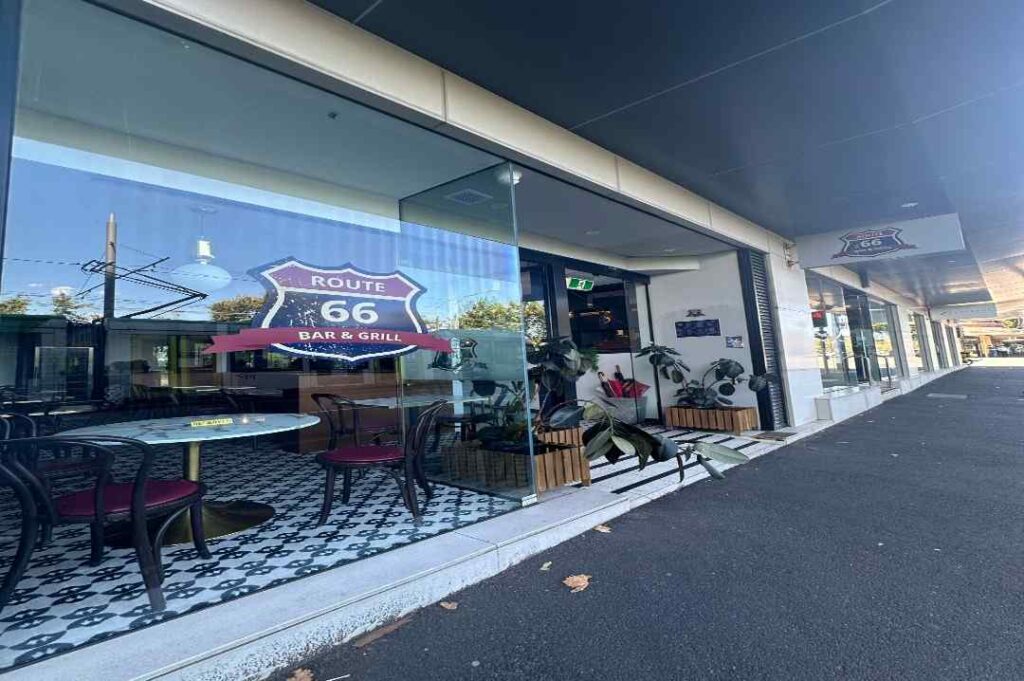 Street view of Route 66 Bar & Grill at 150 Wellington Parade in East Melbourne, showcasing the modern glass-front entrance, indoor seating, and signature Route 66 signage for this American-style restaurant and bar serving burgers, steaks, and classic drinks.